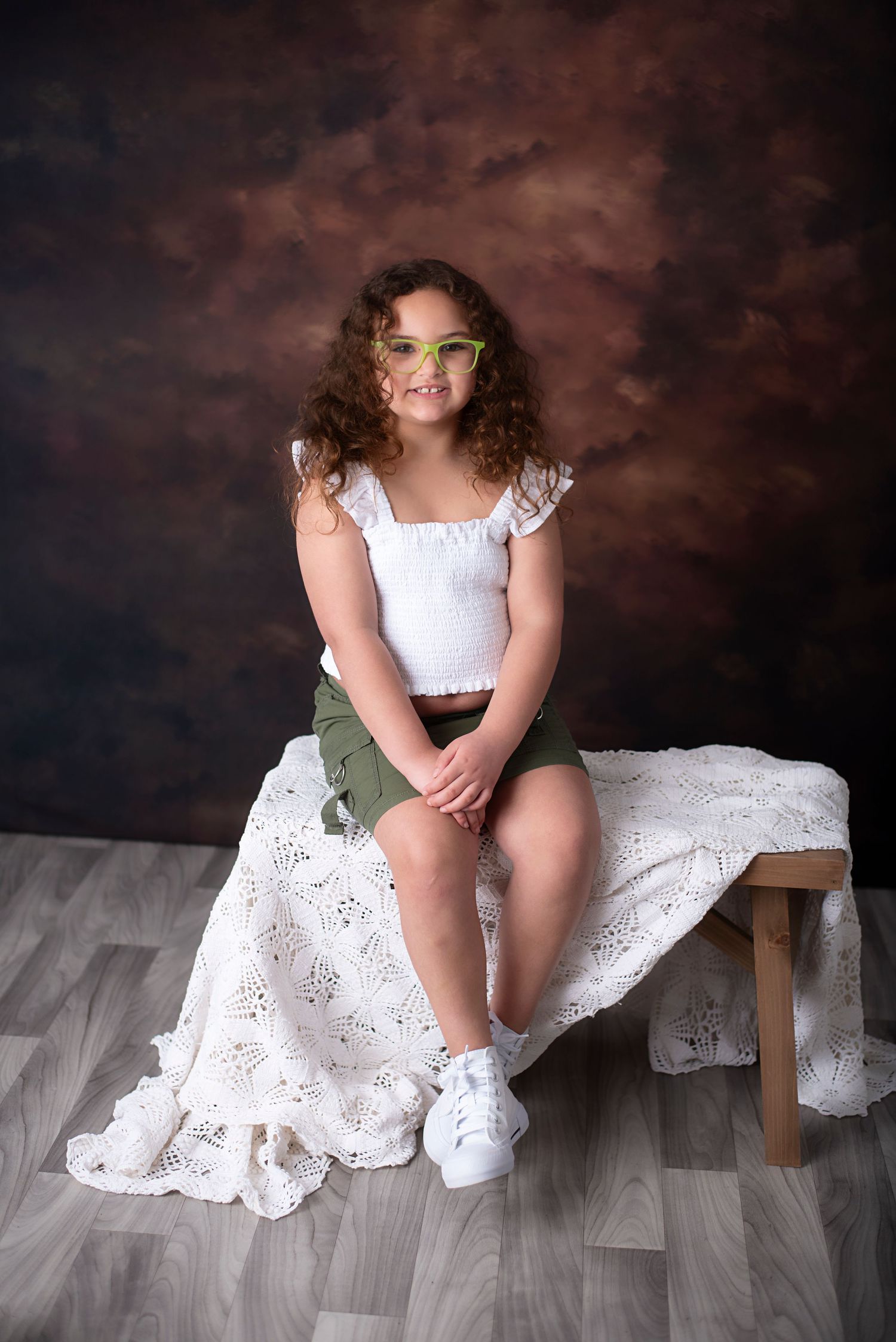 A person in a white eyelet lace dress poses artistically on a wooden bench against a dark studio backdrop.