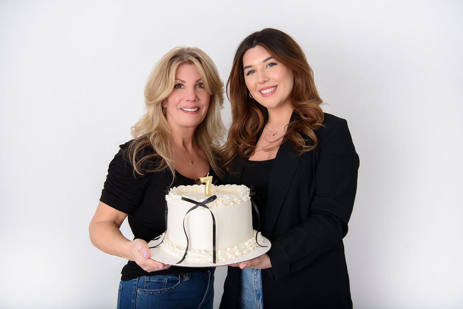 Two people in black outfits hold and smile at a white cake with frosting in a bright studio setting.