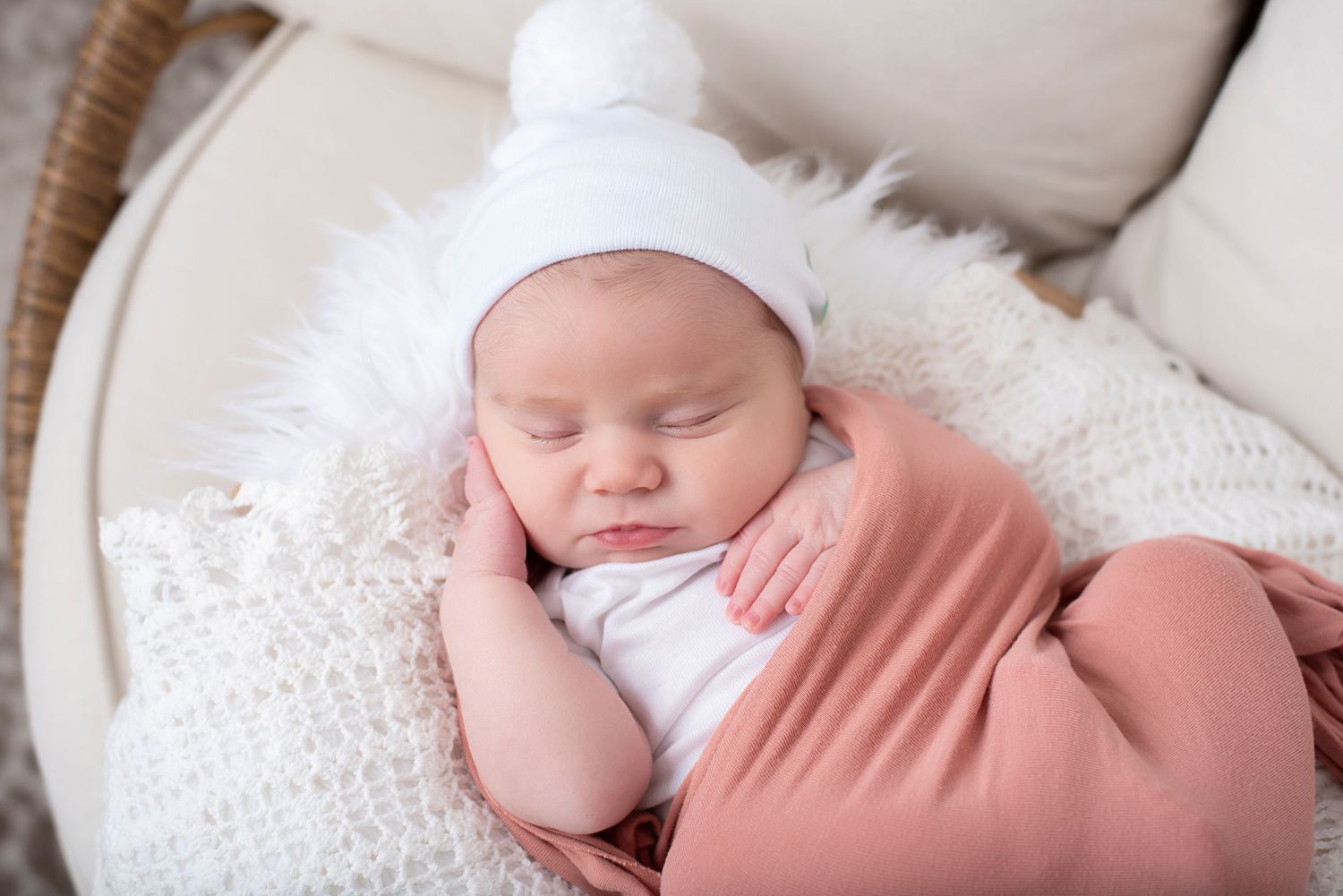 A sleeping newborn in a white knit hat and soft peach wrap rests peacefully on white textured blankets.