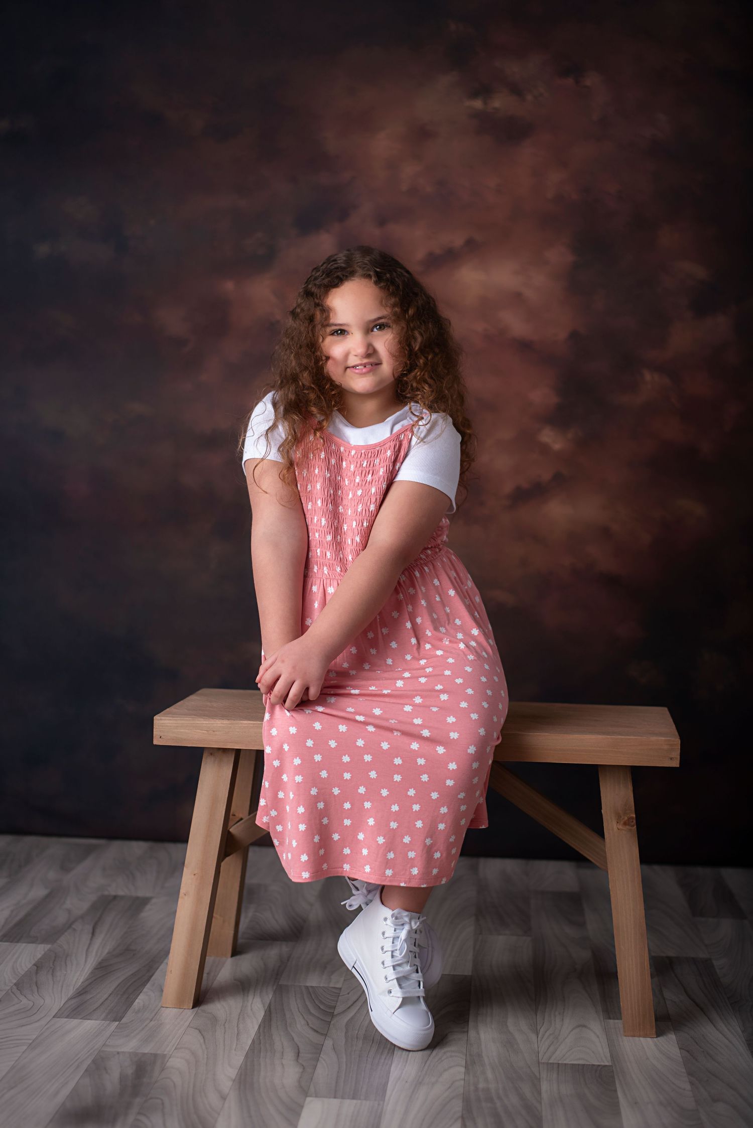 Little girl in polka dot pink dress and white shoes sitting on wooden bench for portrait session.