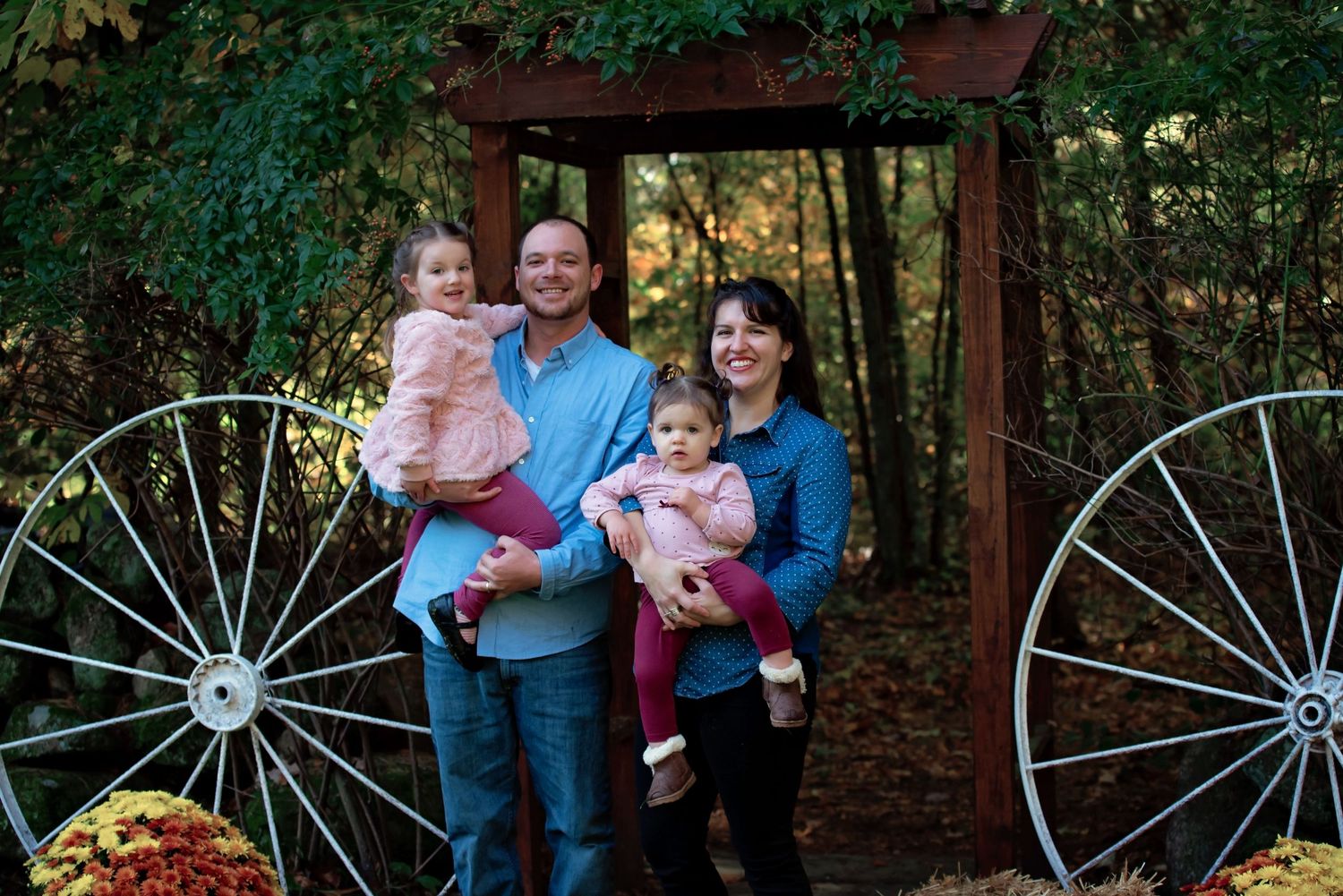 Family portrait with wagon wheels and autumn decorations in a wooded outdoor setting.