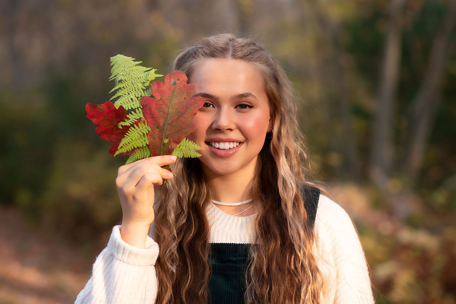 A young person in a white sweater holds red autumn leaves and a fern branch outdoors on a sunny day. Rhode Island Photographer