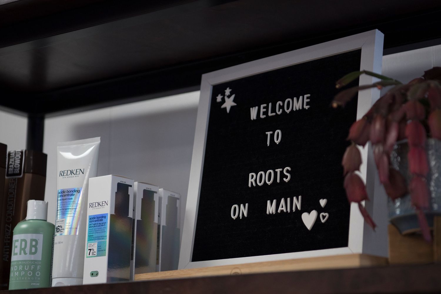 A black letter board sign reading 'Welcome to Roots on Main' mounted on a white wall with blurred products in foreground.