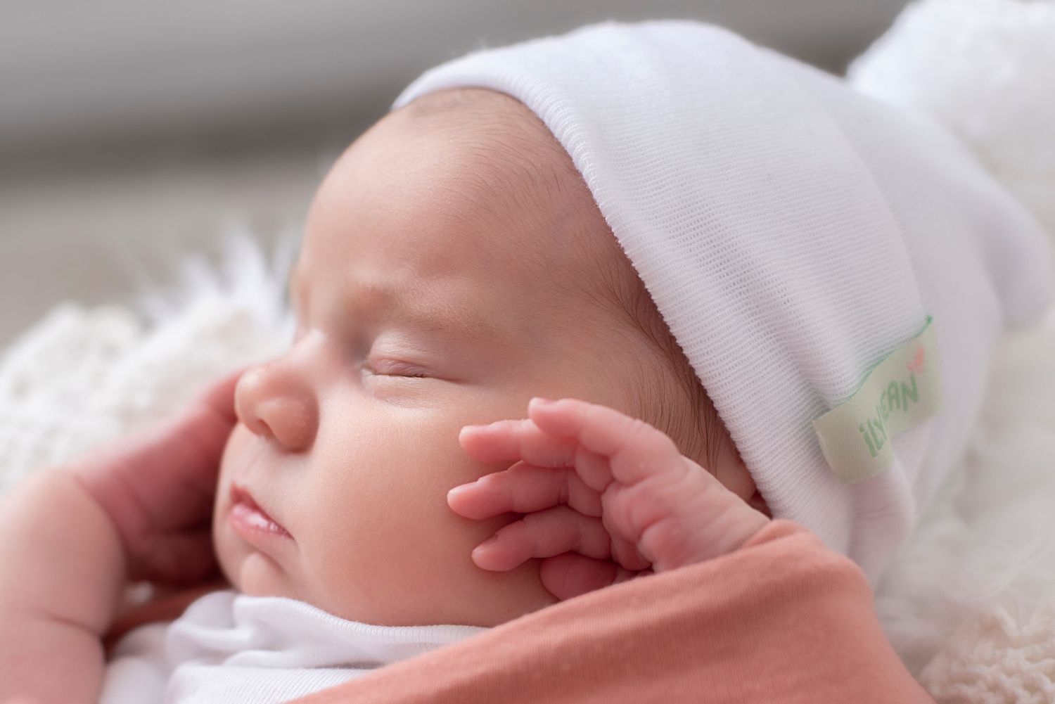 A sleeping newborn baby wearing a white hat rests peacefully against a soft white blanket.