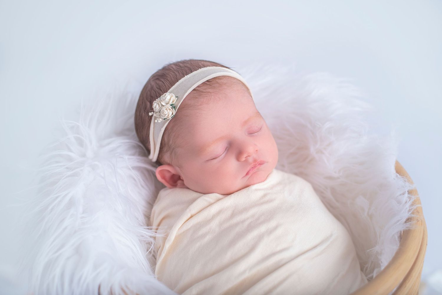 Peaceful newborn baby wrapped in white swaddle sleeps on fluffy white blanket wearing delicate floral headband.
