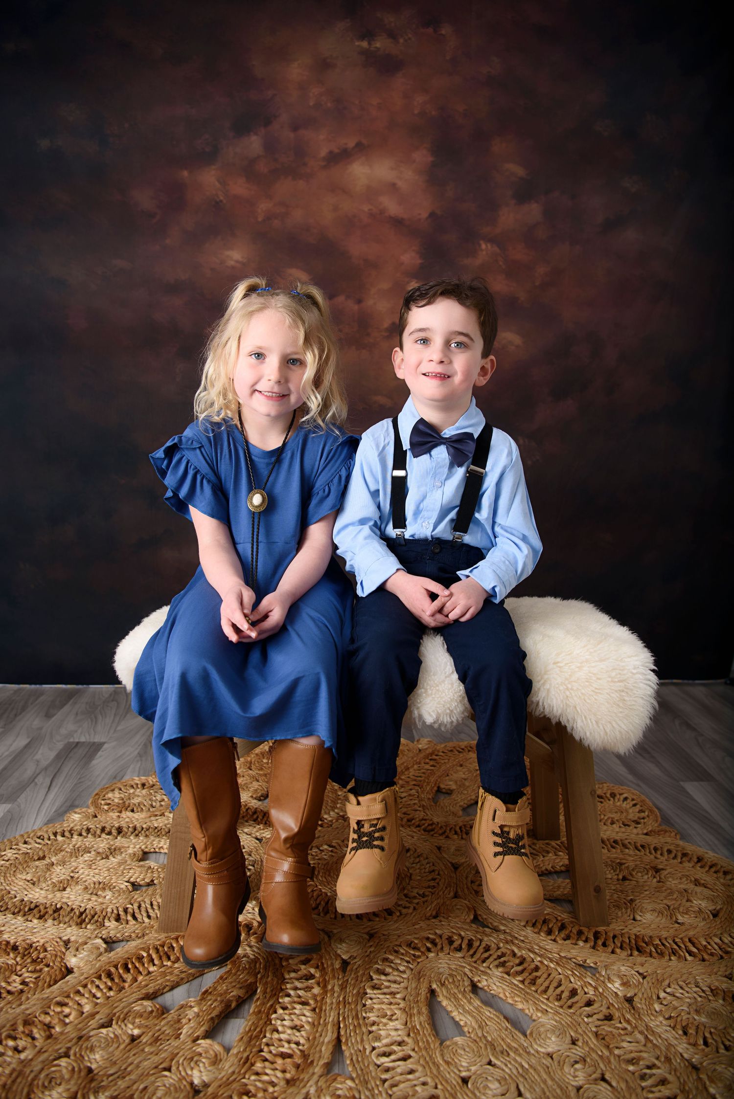 Two children in formal attire sitting together on a furry rug against a dark backdrop wearing matching boots.