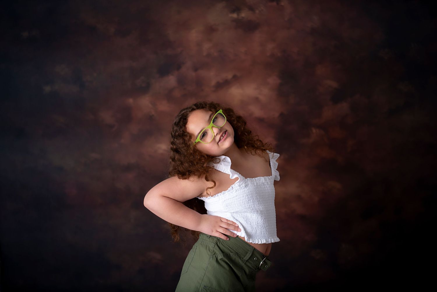 A young dancer in a white top and olive skirt strikes a graceful pose against a dark brown backdrop.