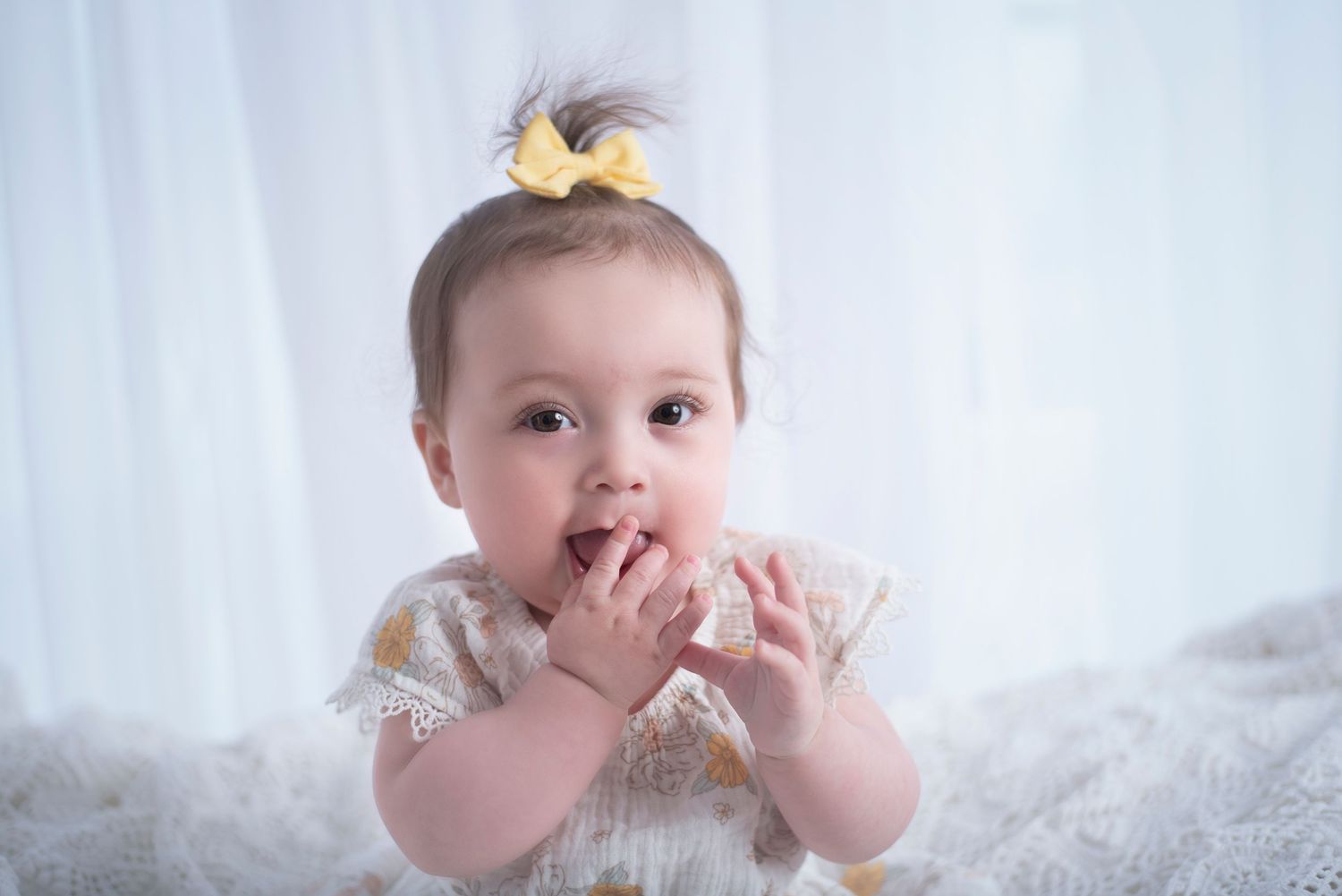 Sweet baby in white dress with yellow bow lying on fluffy white blanket with hands near mouth.