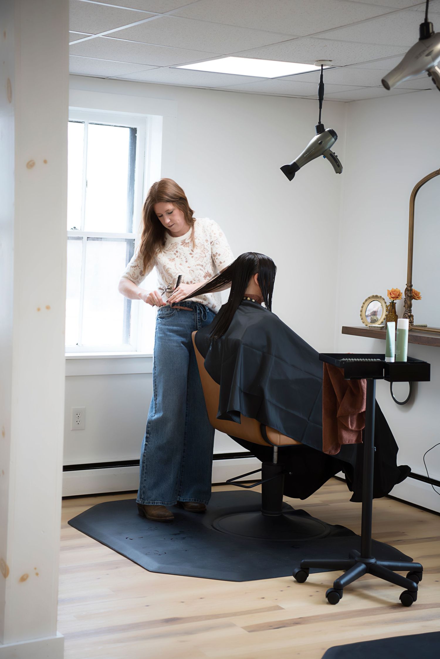 A stylist works on a client's hair in a bright, minimalist salon setting with hardwood floors and white walls.