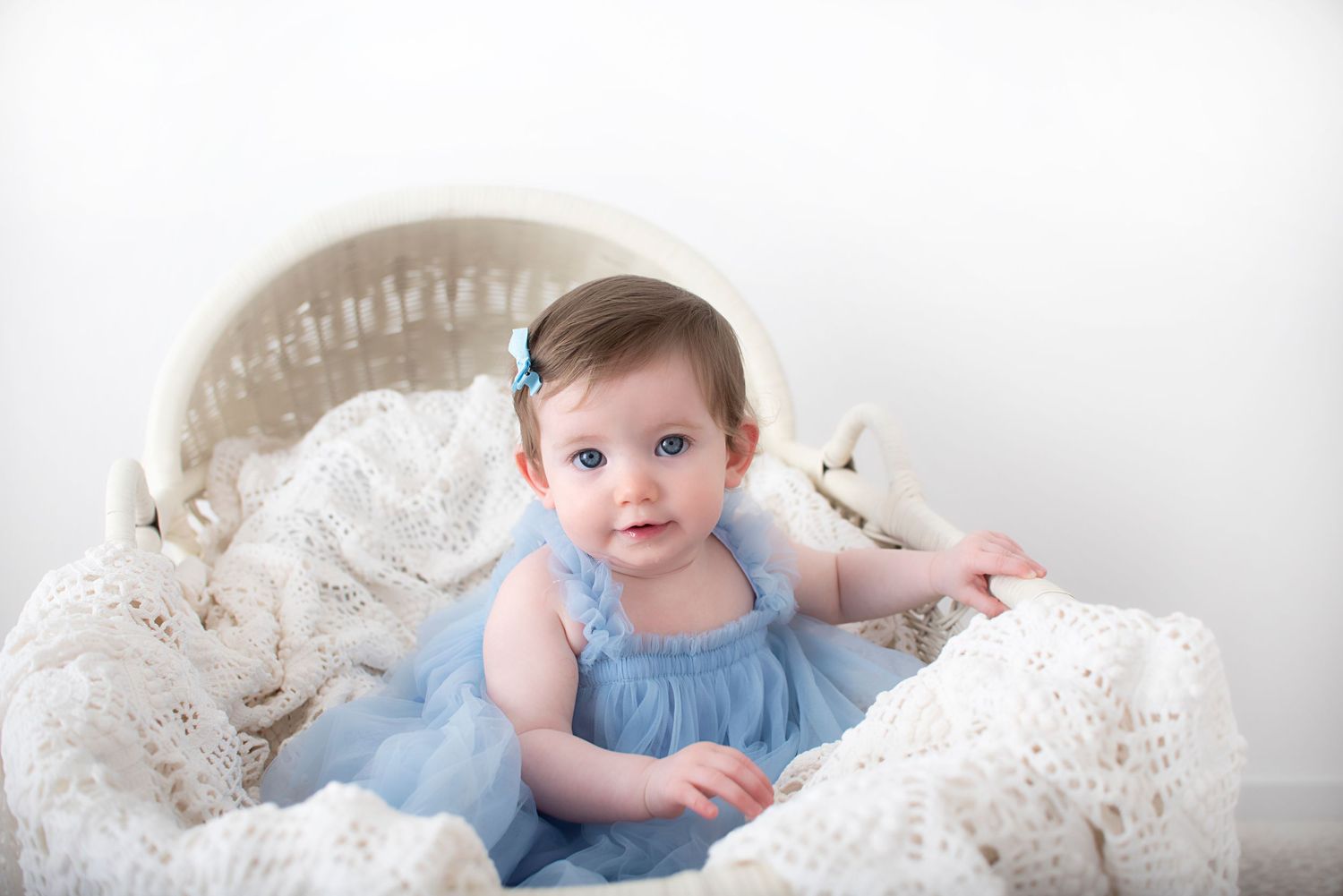 Baby in light blue dress sits on white lace blanket during portrait session.