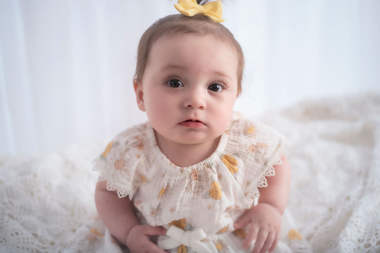 Baby wearing a delicate white and yellow lace dress sits on soft white fabric with a yellow bow in their hair.