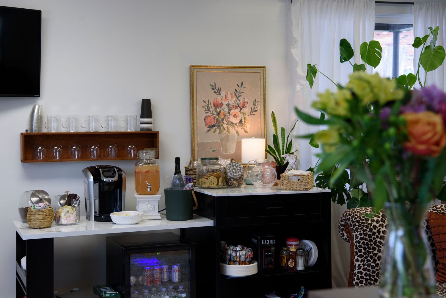 A cozy coffee station corner with artwork, storage shelves, and fresh flowers in a vase.