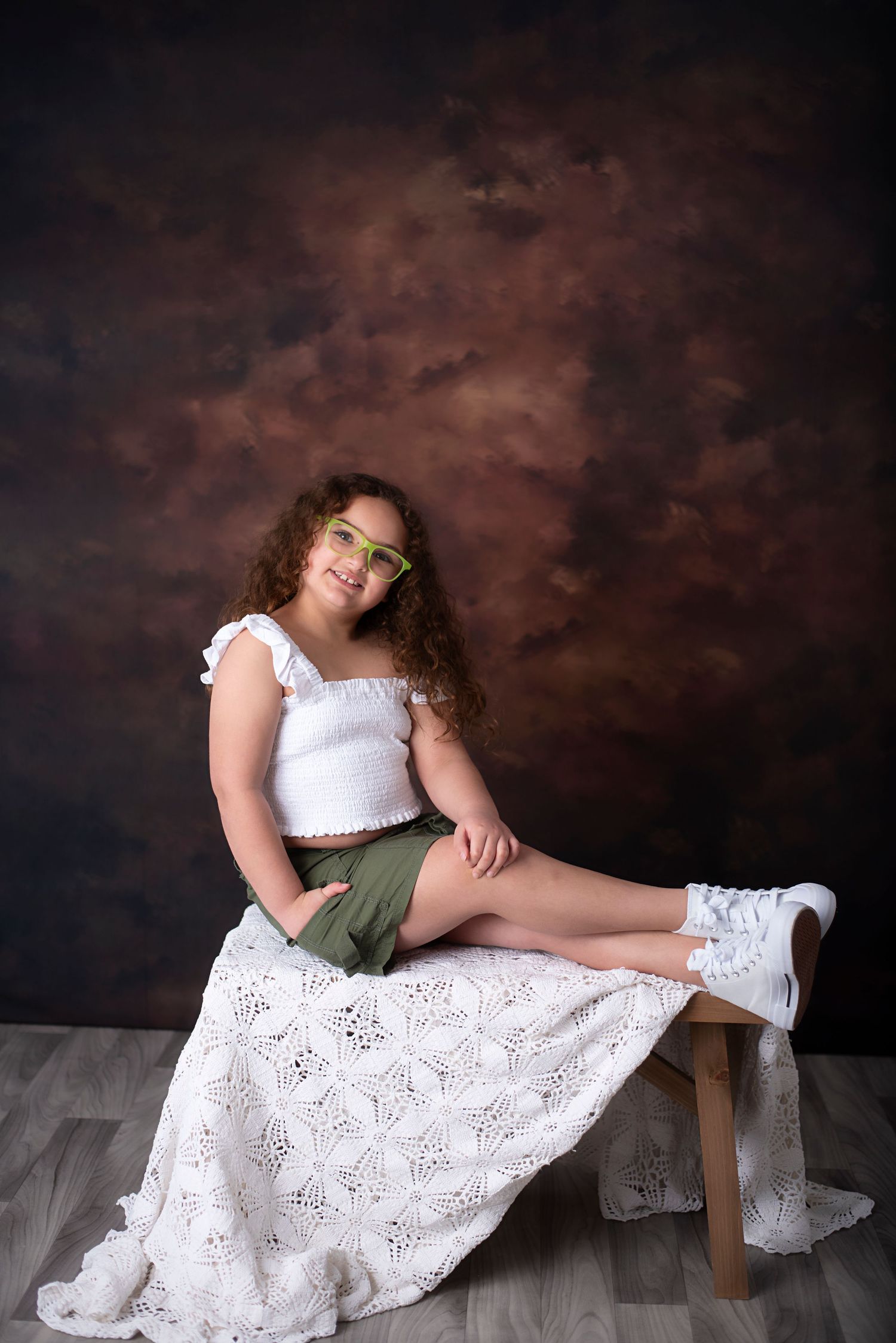 Artistic portrait of someone wearing a white lace crop top and skirt with curly hair against a moody brown backdrop.