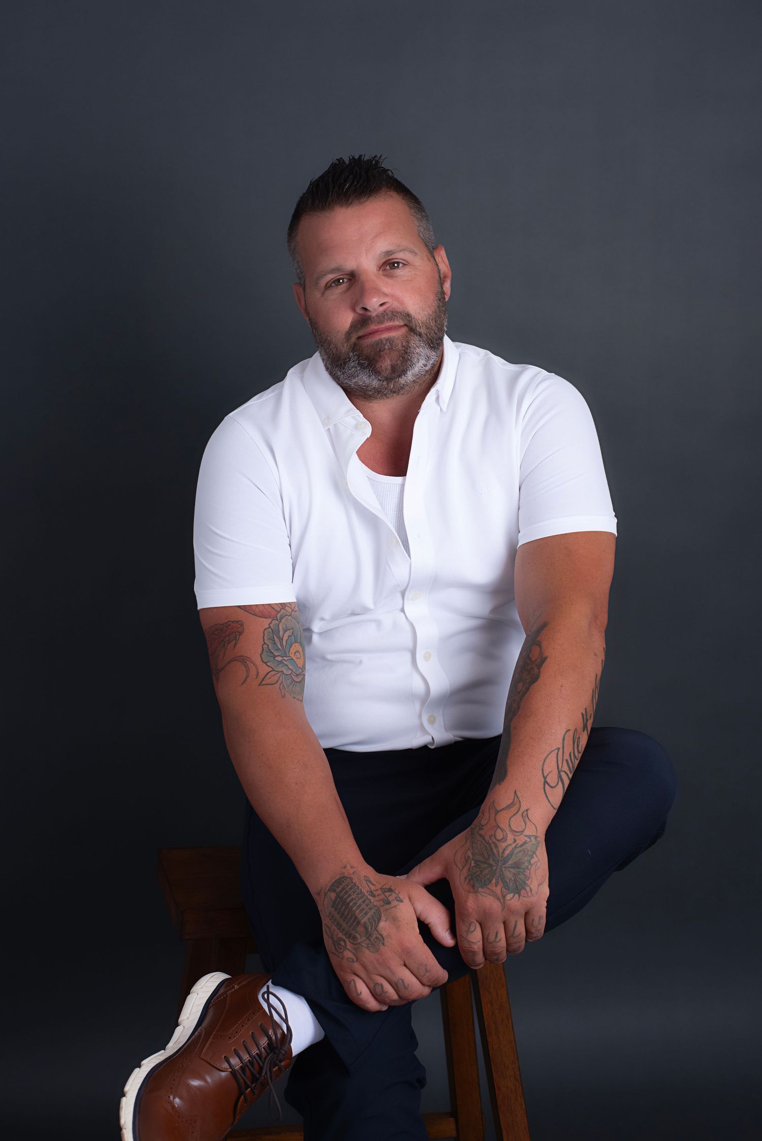 Muscular man in white t-shirt with tattoos sits on wooden stool against dark background in studio portraits.