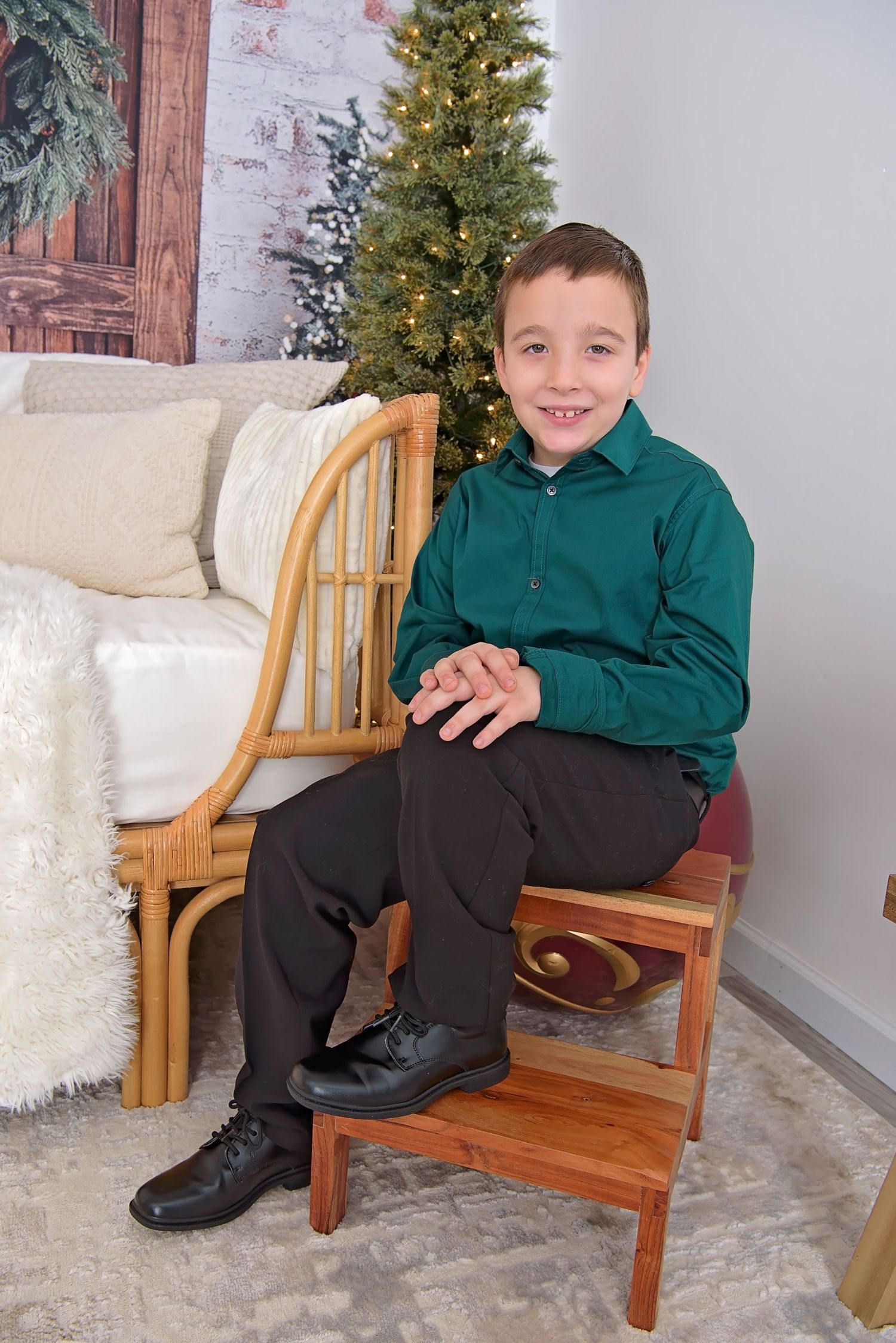 Child in green shirt and black pants sits on wooden bench with Christmas tree and winter decor in background.