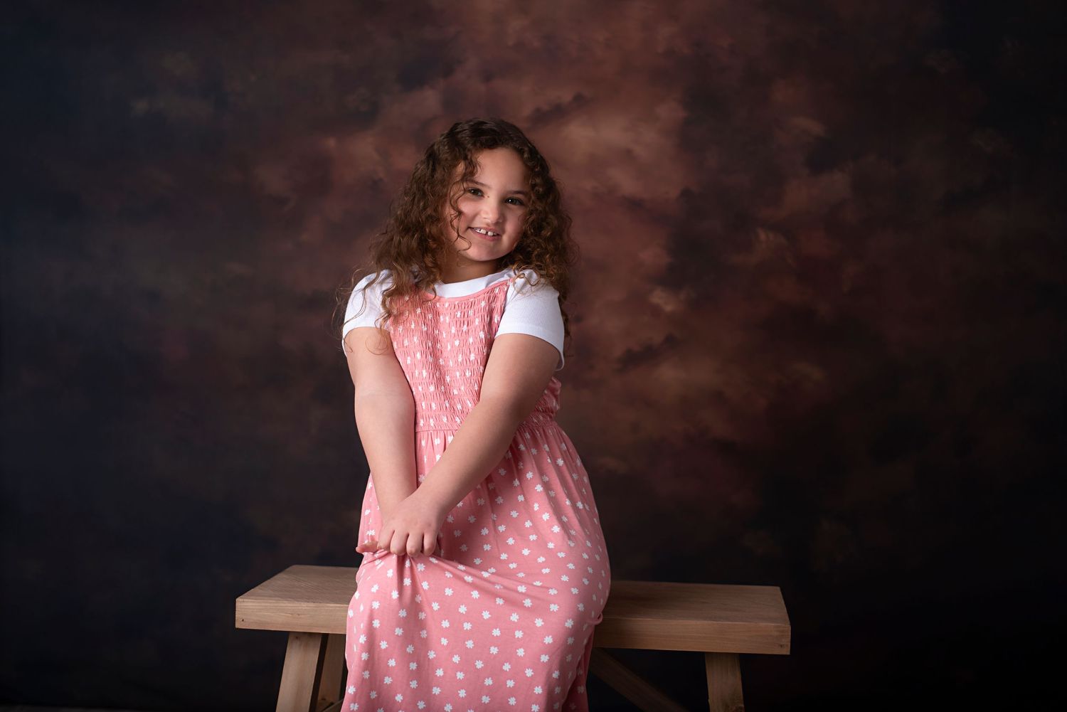 Figure in a pink polka dot dress and white top sits on wooden bench against dark studio background.