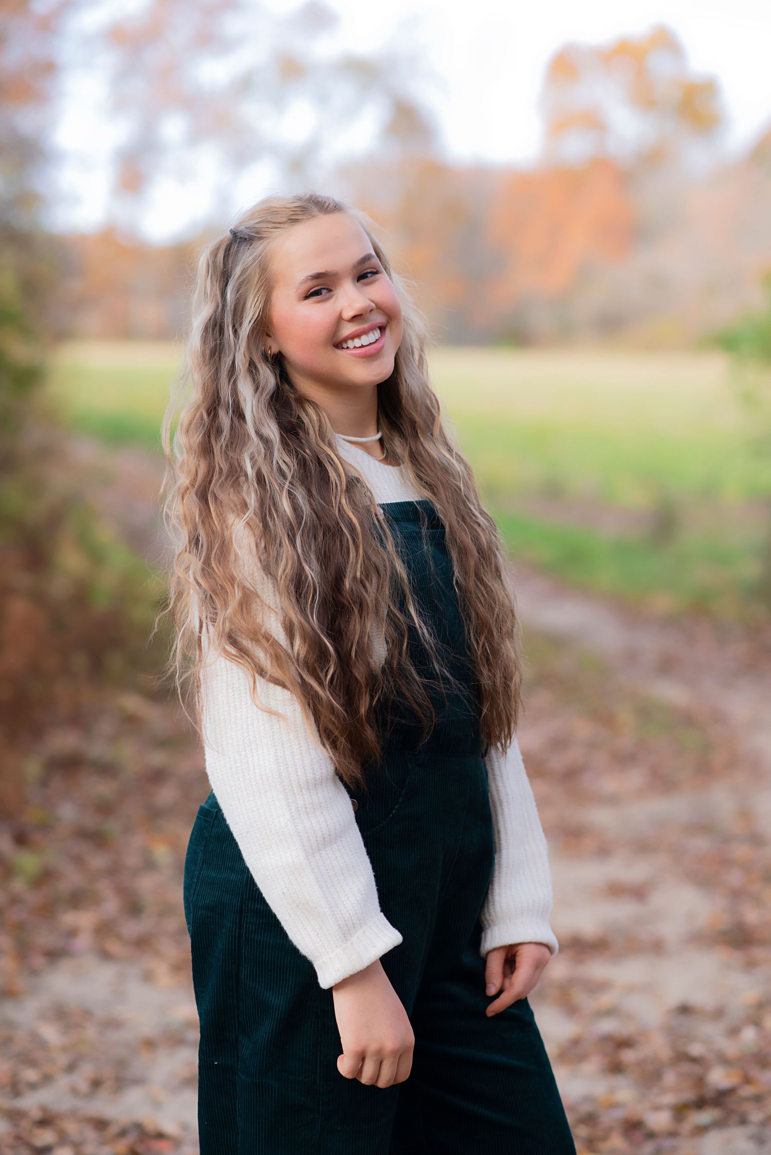 Smiling person with long wavy blonde hair wearing a dark dress and white sweater poses outdoors in autumn.