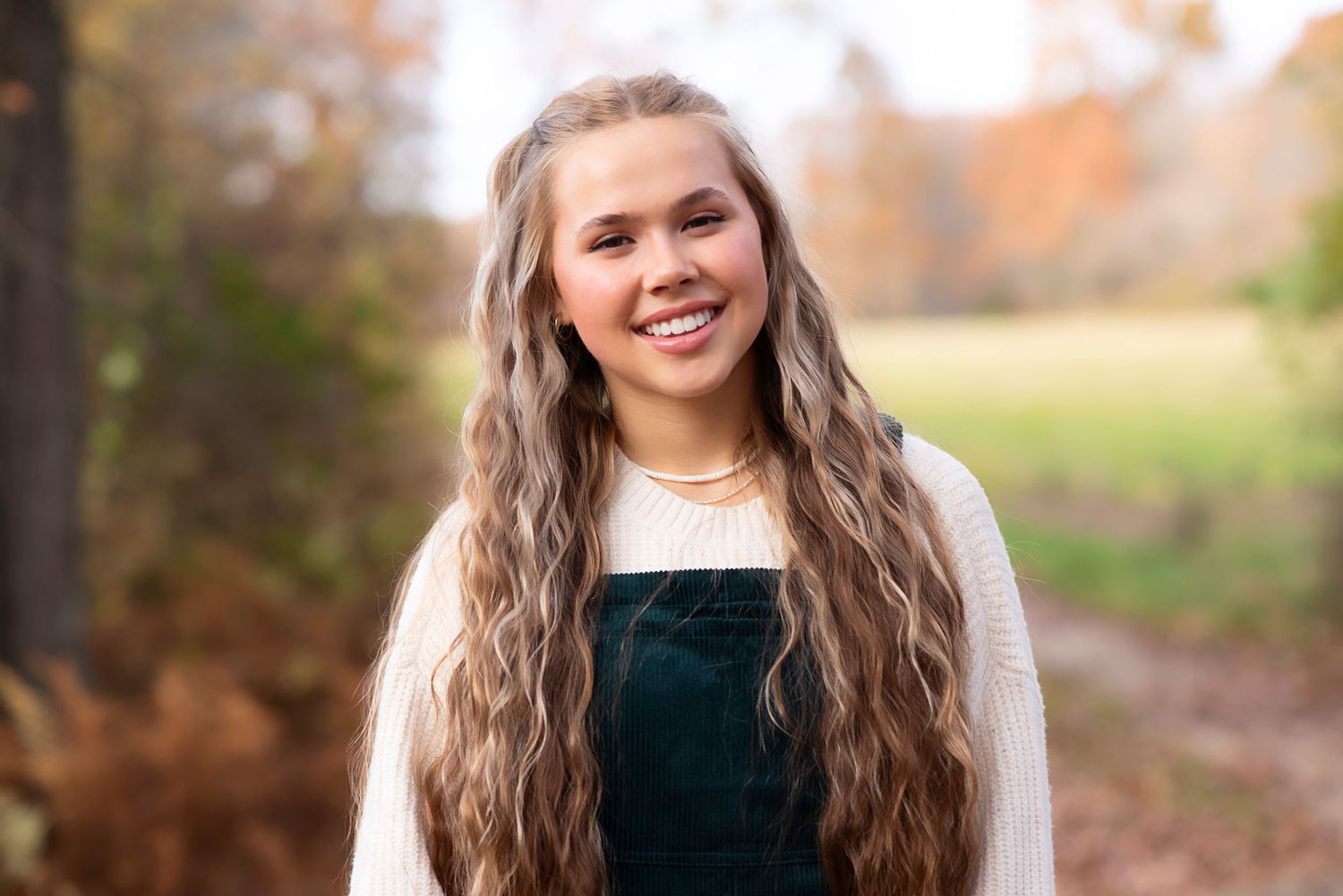 Someone wearing a dark green velvet overall dress and cream sweater poses outdoors during autumn.