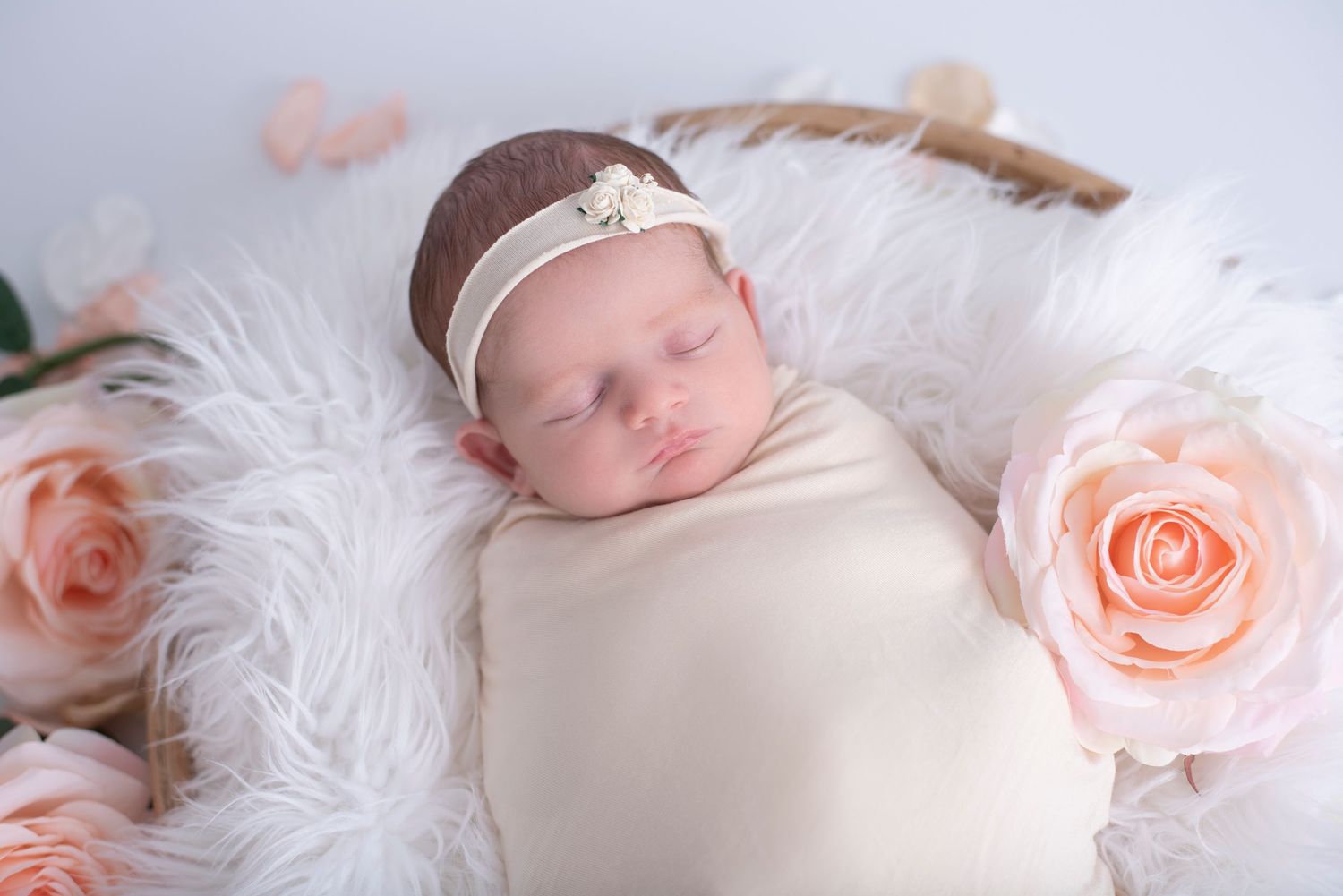 Peacefully sleeping newborn swaddled in white on soft white fur blanket with delicate peach roses.