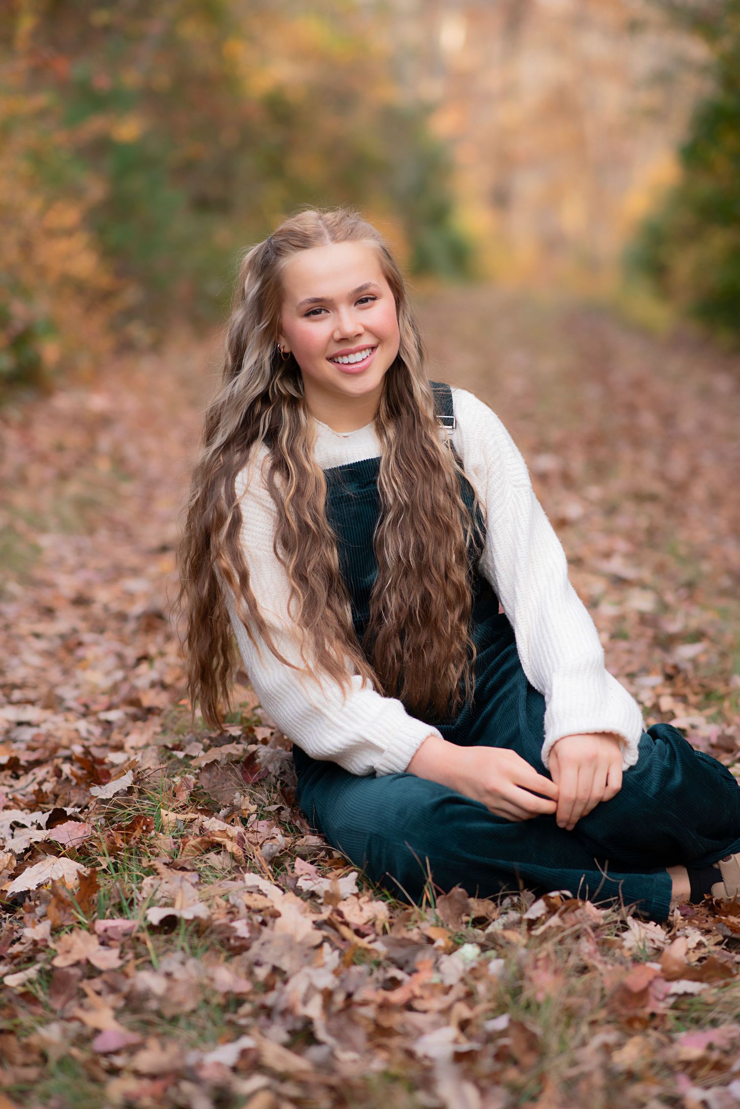 A person in a white sweater and dark dress sits smiling among fallen autumn leaves on a forest path. Rhode Island Photographer