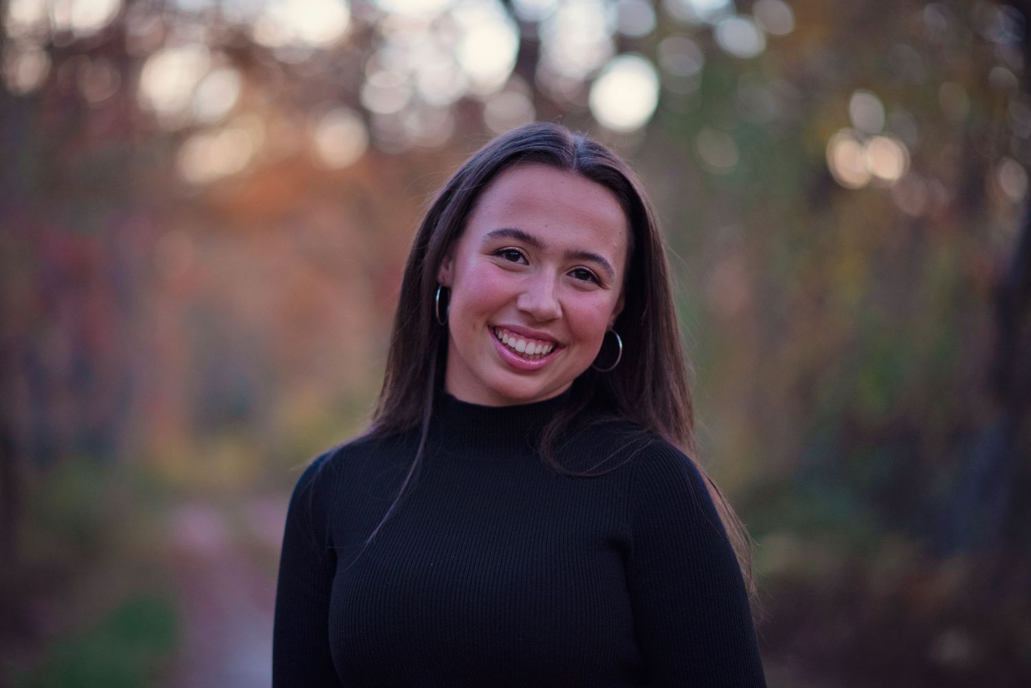 A smiling figure wearing a black turtleneck sweater poses outdoors with autumn foliage creating a soft, blurred background.