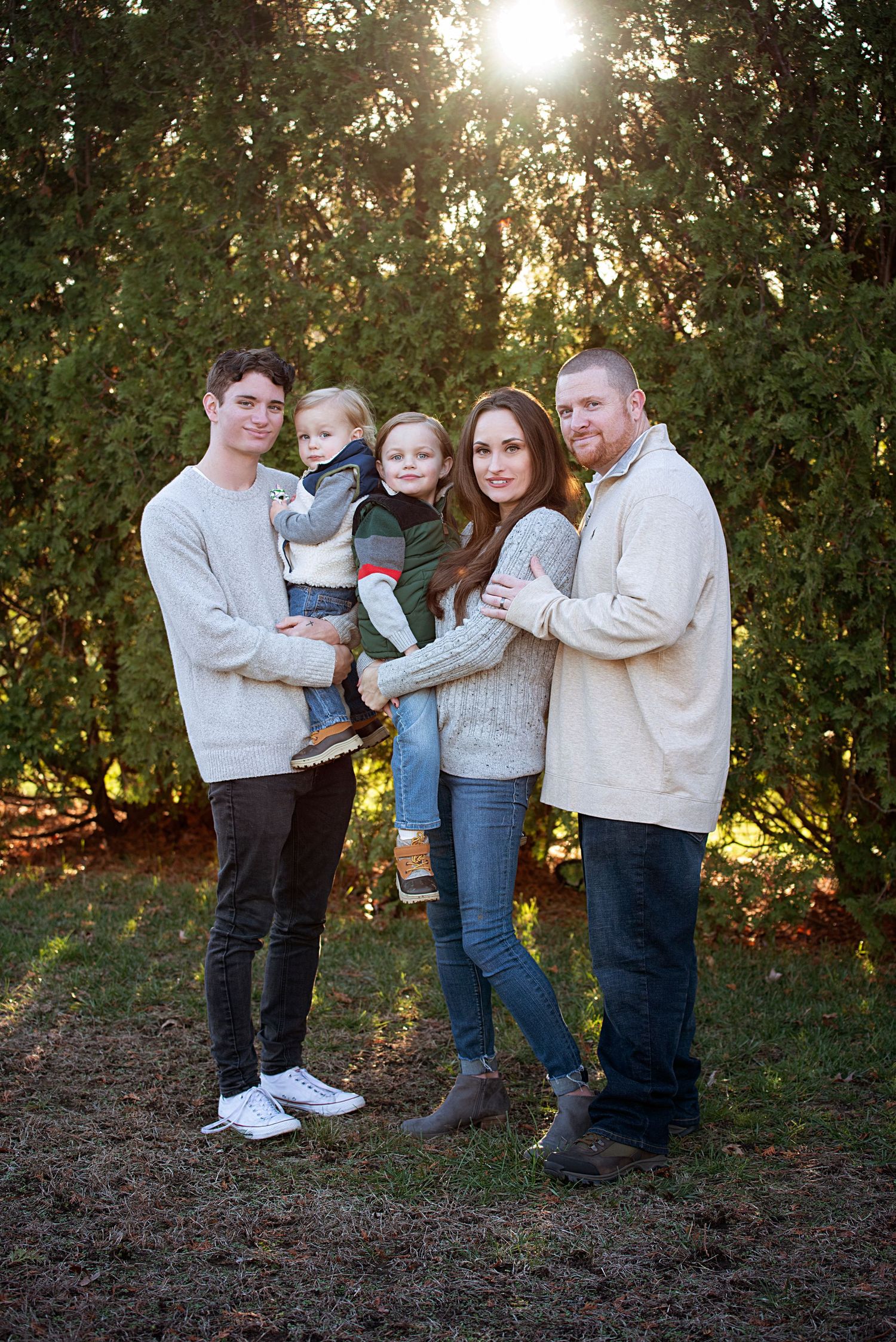Group in matching cream sweaters poses together in a forest setting at golden hour with sunlight peeking through trees.