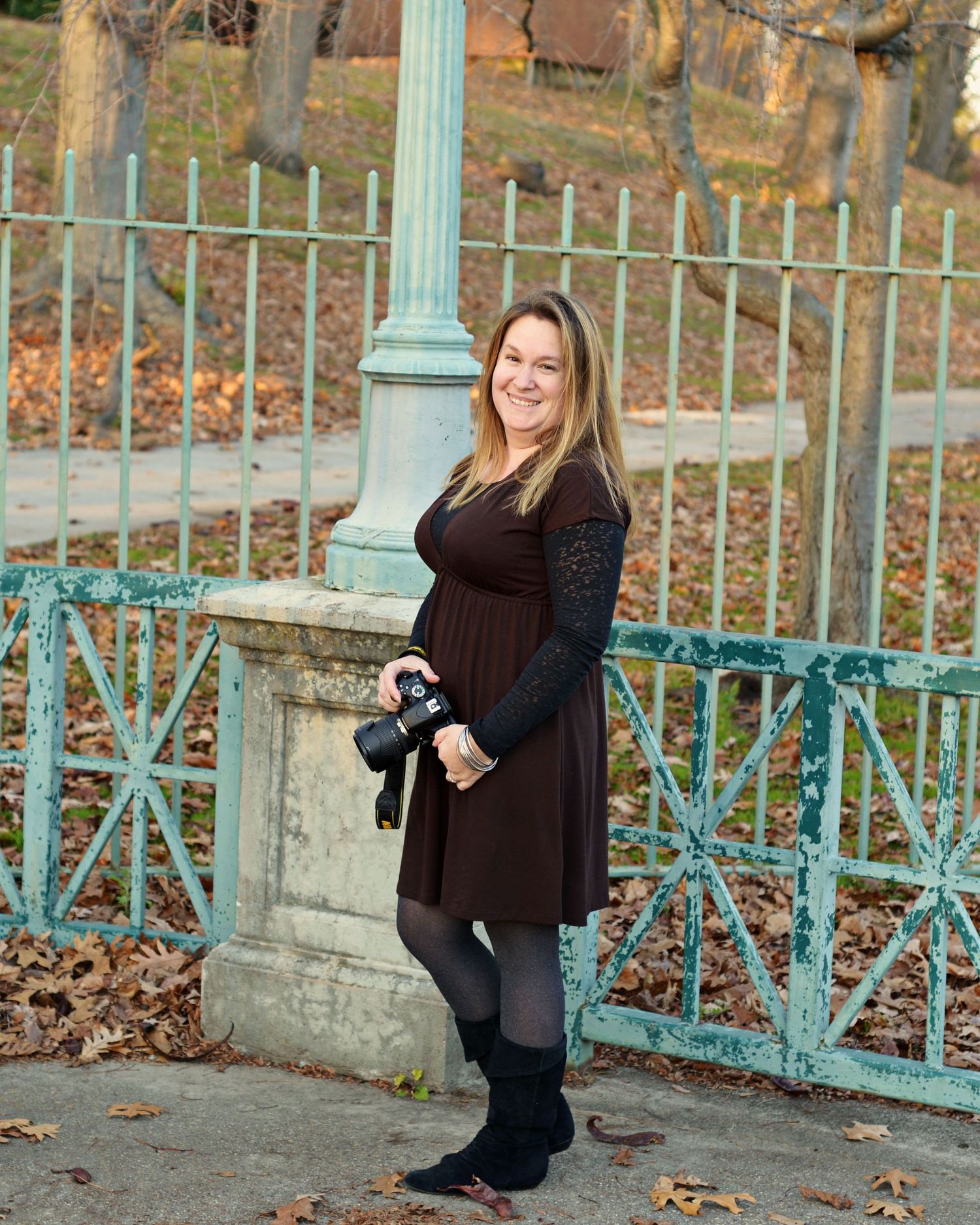 Exeter, RI Photographer poses against a decorative turquoise iron fence with autumn leaves scattered on the ground Roger Williams Park.