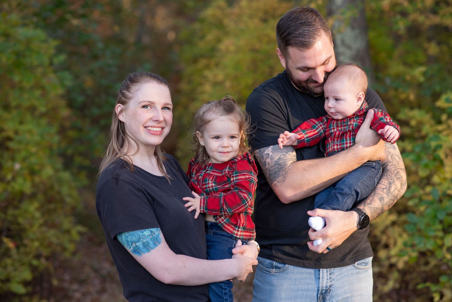 Family embraces while wearing matching red plaid and black clothing during fall photo session in wooded area.