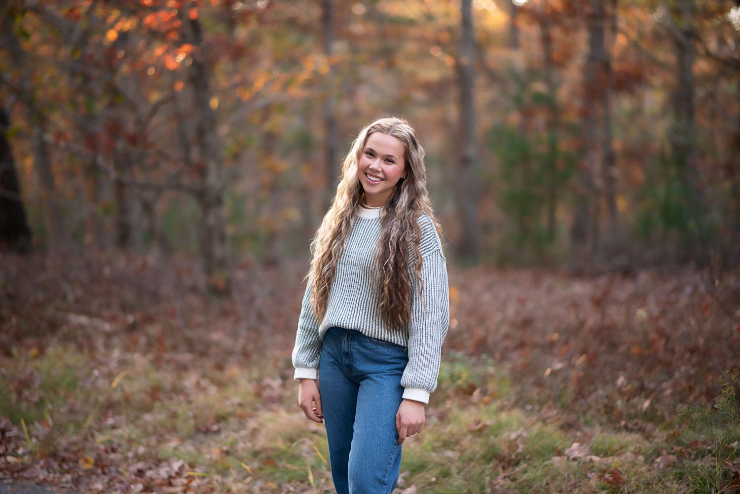 Smiling figure wearing a cozy gray sweater and jeans stands in autumn woods surrounded by fallen leaves. Rhode Island Photographer