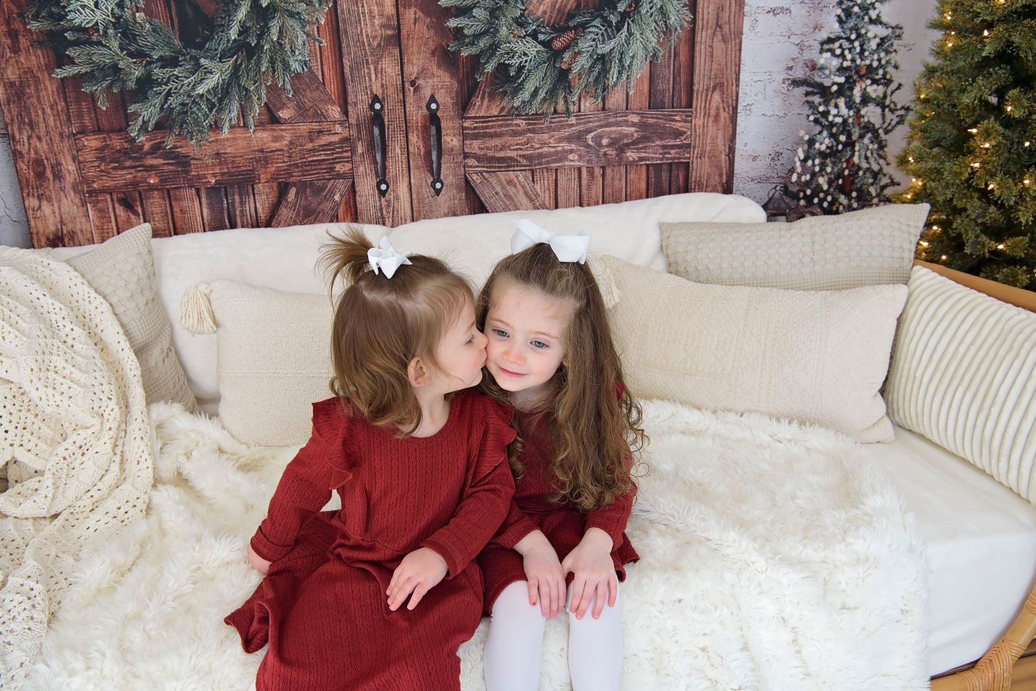 Two young children in holiday attire share a sweet moment on a cozy white couch with Christmas decor in background.