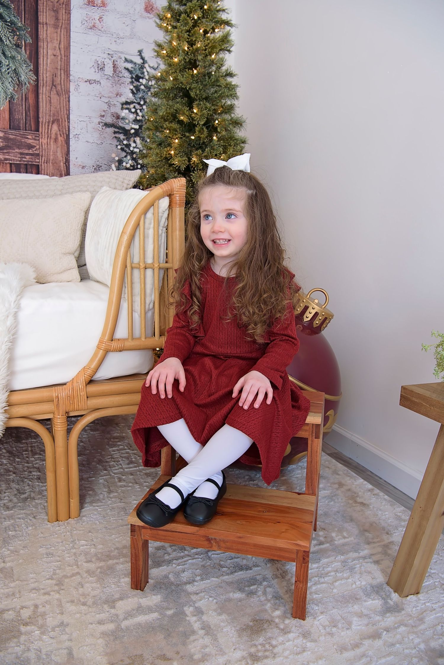 Child in burgundy dress sitting on wooden bench with festive Christmas tree in background.