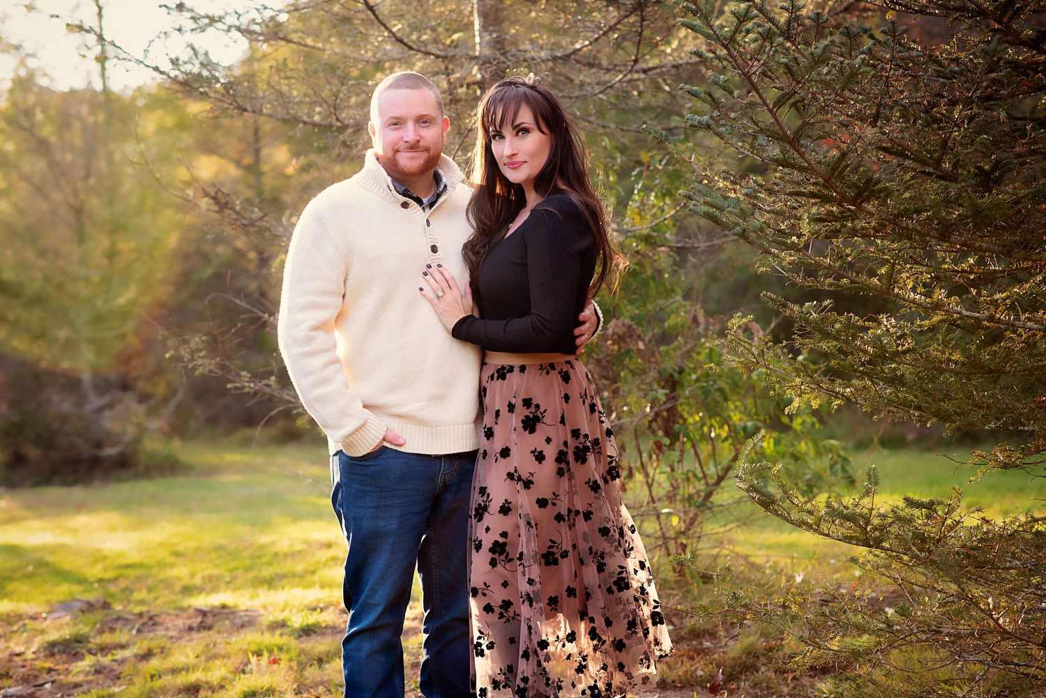 A couple poses together in a wooded outdoor setting during autumn with soft natural lighting and fall foliage.