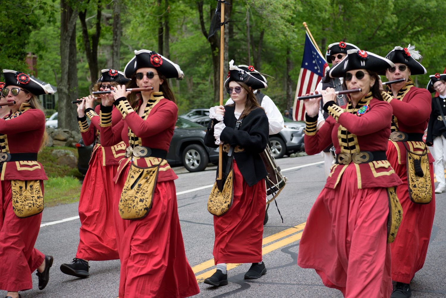 Colonial reenactment fife and drum corps in red period uniforms marching in parade with American flag visible.