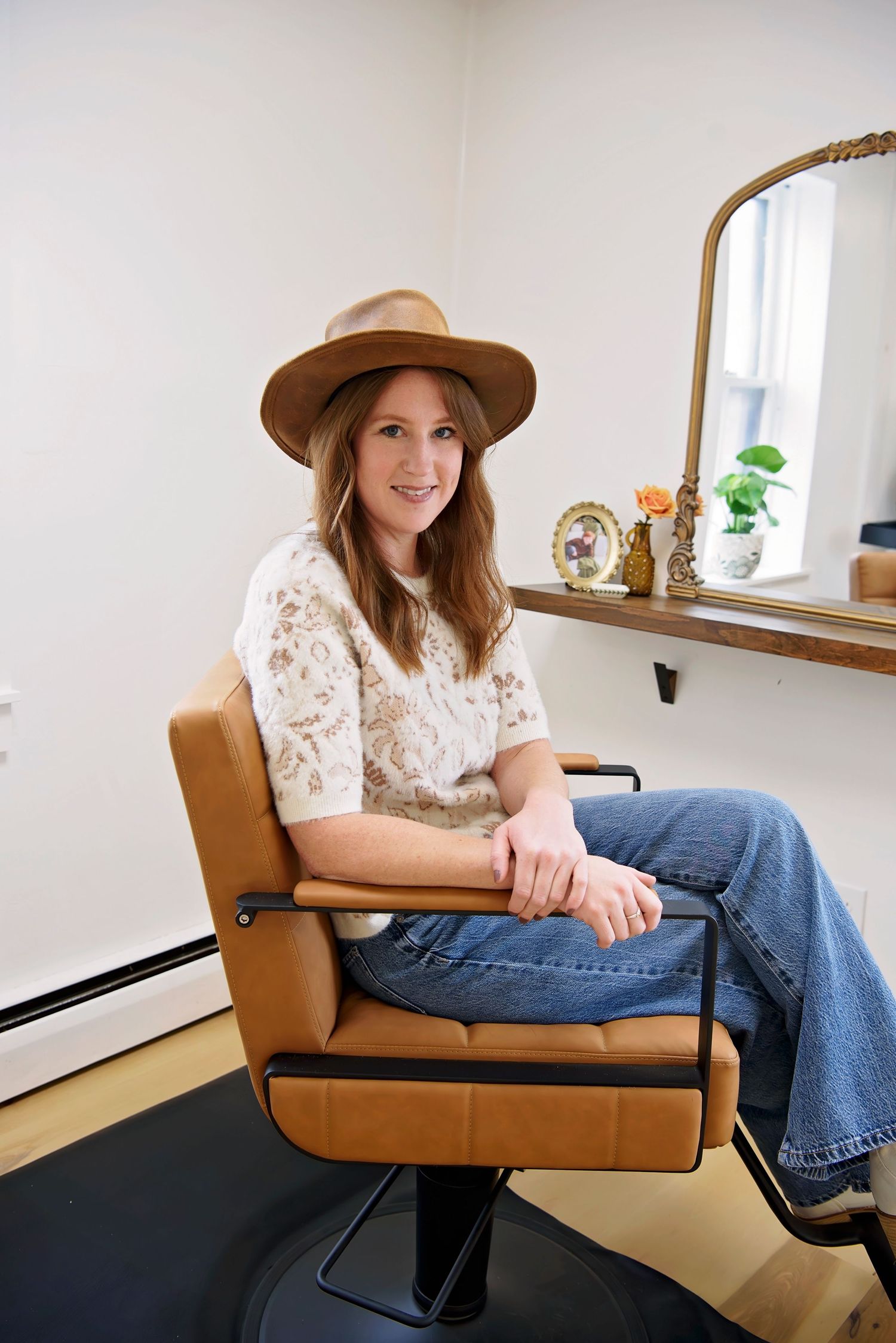 A person wearing a brown hat and white lace top sits in a vintage tan leather salon chair next to a decorative mirror.