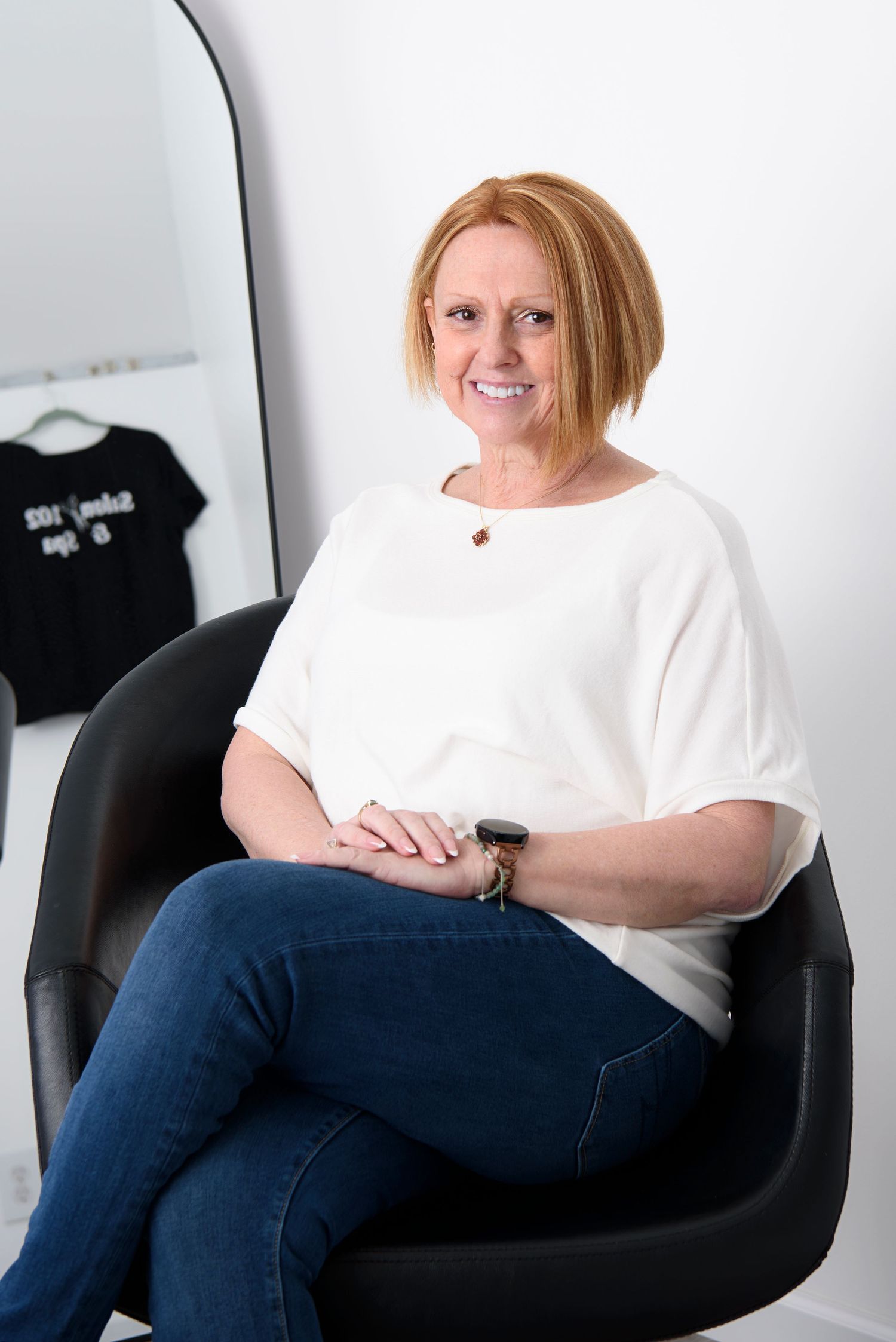 Person in white t-shirt and jeans sits casually on black chair against white wall with dark clothing hanging behind.
