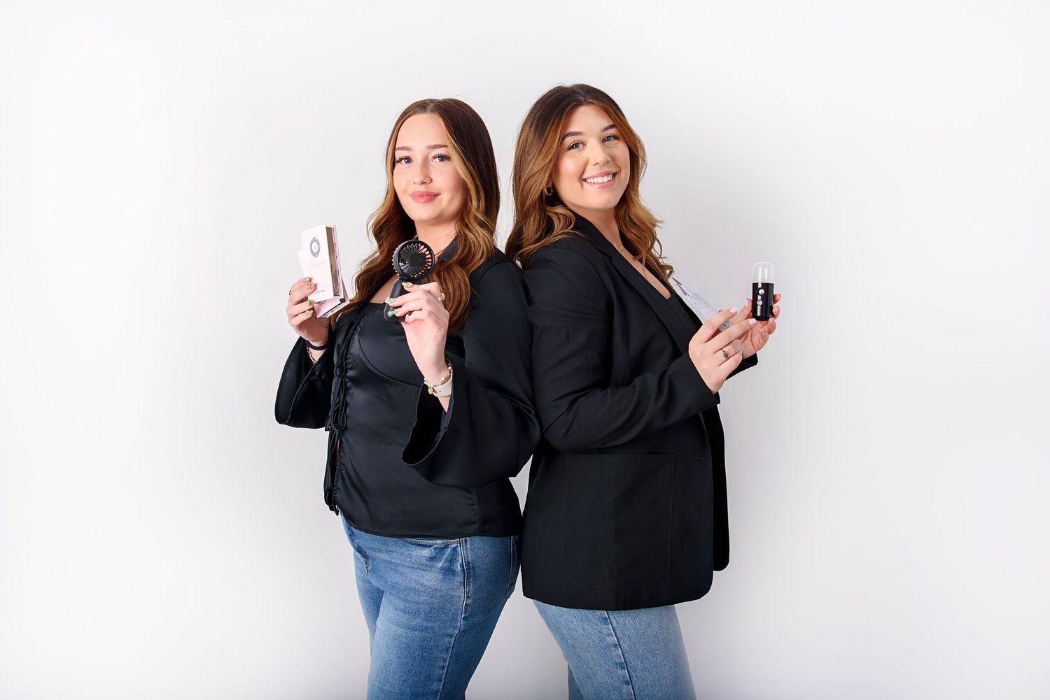 Two women in black sweaters and jeans pose together while smiling and holding drinks against a white background.