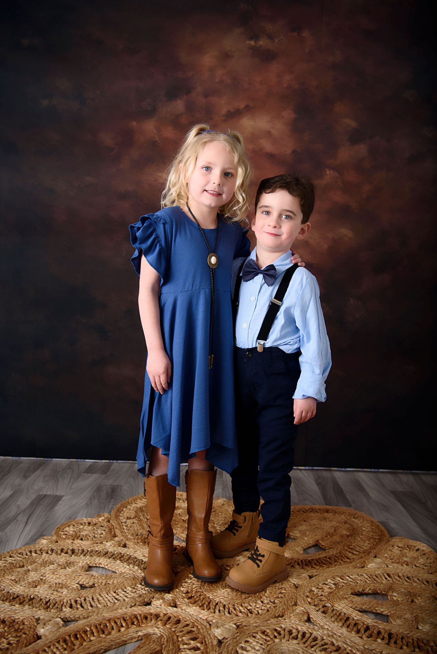 Two children in coordinated formal outfits pose on a patterned rug against a dark dramatic backdrop.