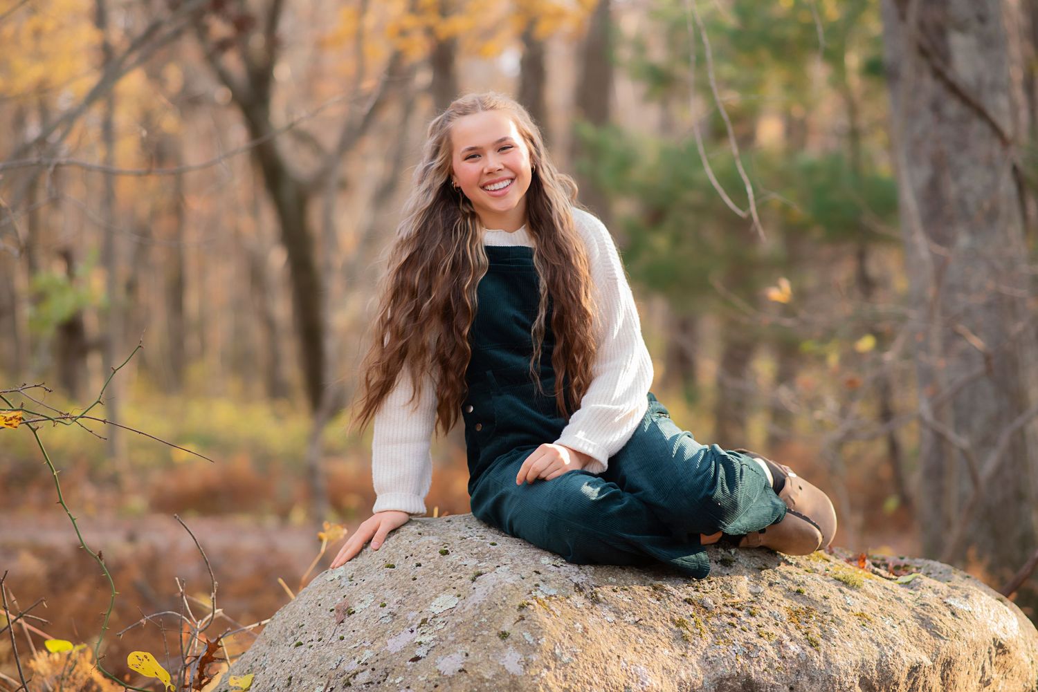 Someone in a green sweater sits atop a large gray boulder in an autumn forest setting during golden hour.