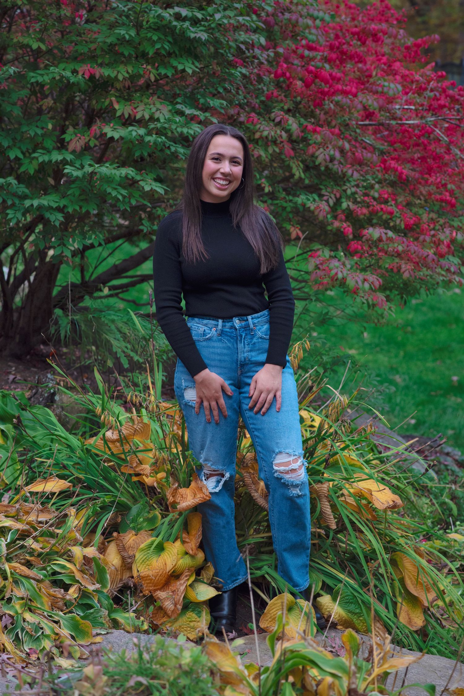 A figure sits in a garden against a backdrop of vibrant red autumn leaves and lush green foliage on a fall day.