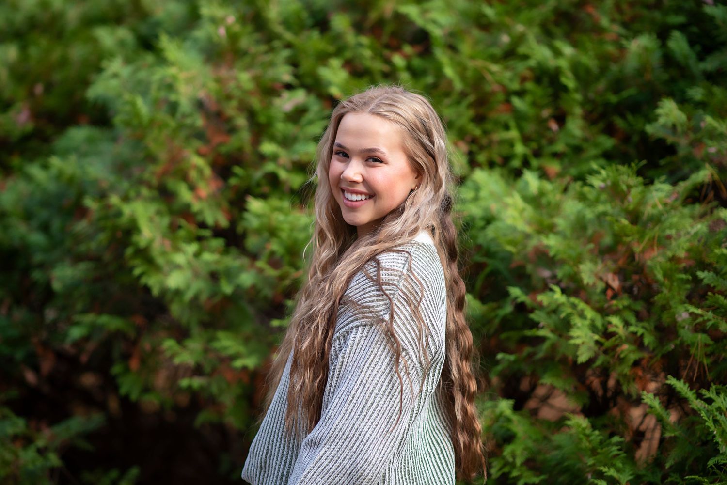 Young person in grey sweater smiling while standing outdoors with green foliage in background. Rhode Island Photographer