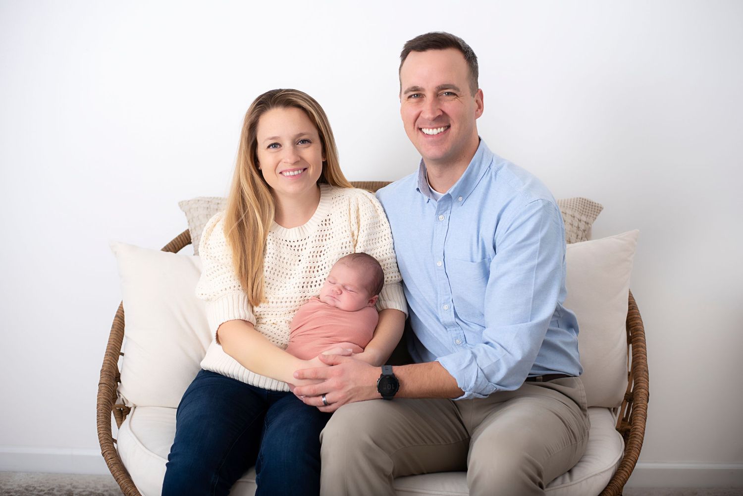 A family sits together on a woven wicker loveseat against a white wall for a portrait session. RI Newborn Photographer