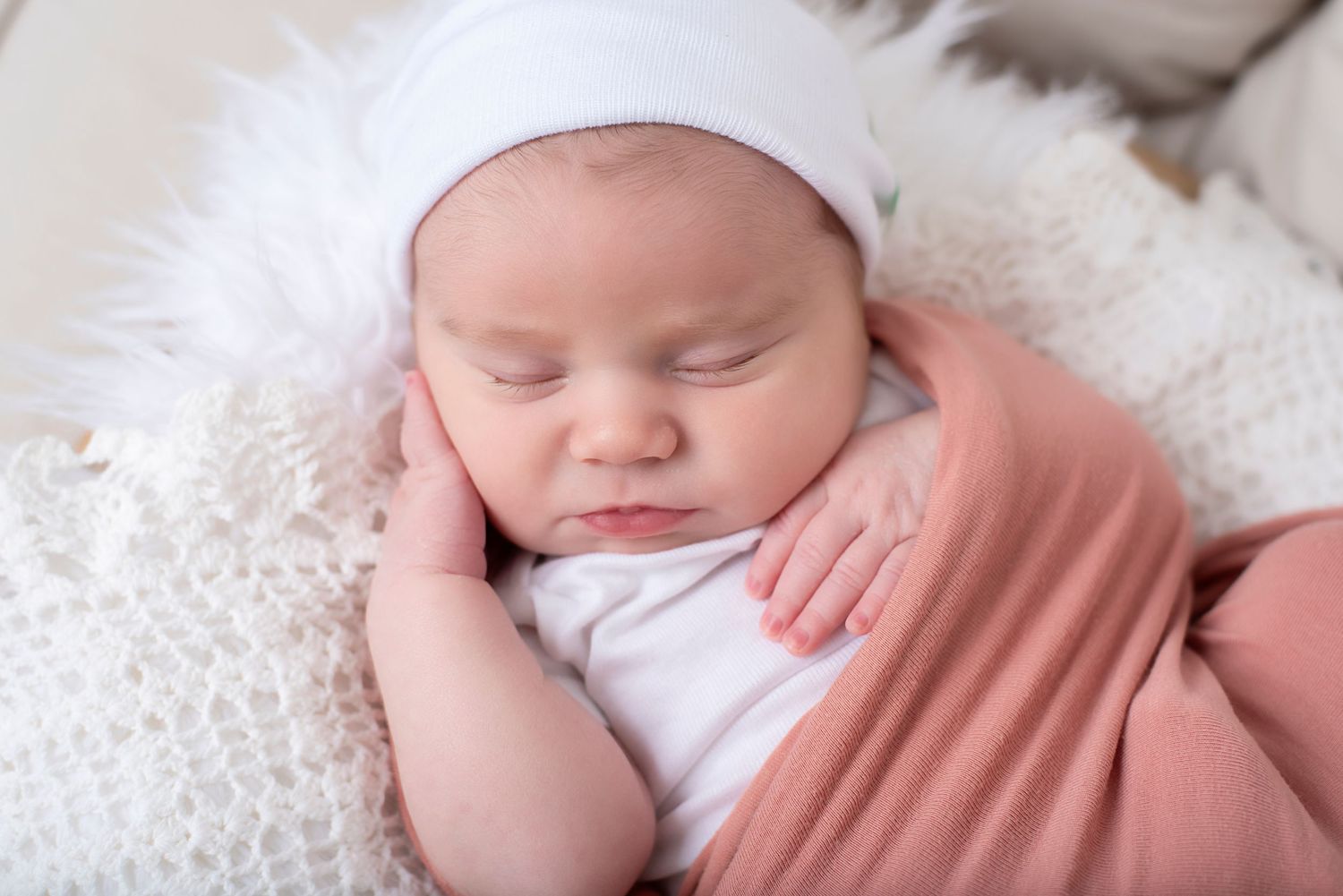Sleeping newborn baby swaddled in soft pink fabric resting on white fluffy blanket during photo session.