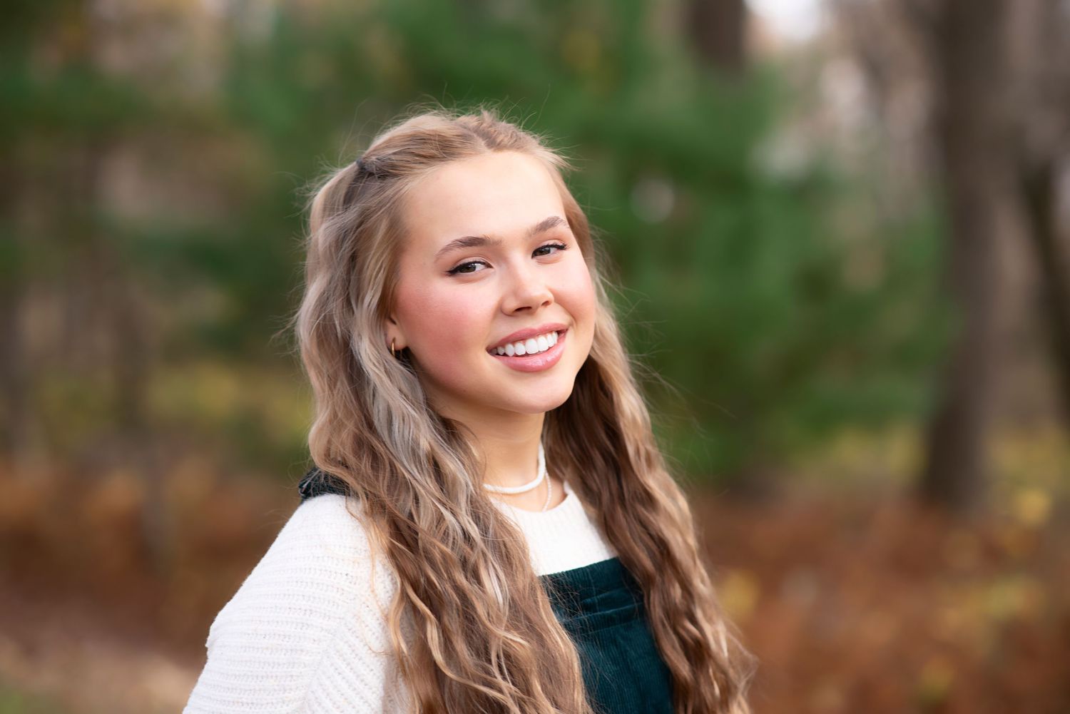 A smiling student with long wavy hair wearing a white sweater outdoors during autumn.