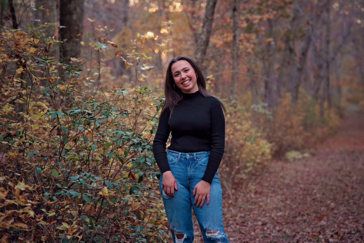 Someone in a black sweater and ripped jeans poses outdoors on a serene autumn path surrounded by fall foliage.