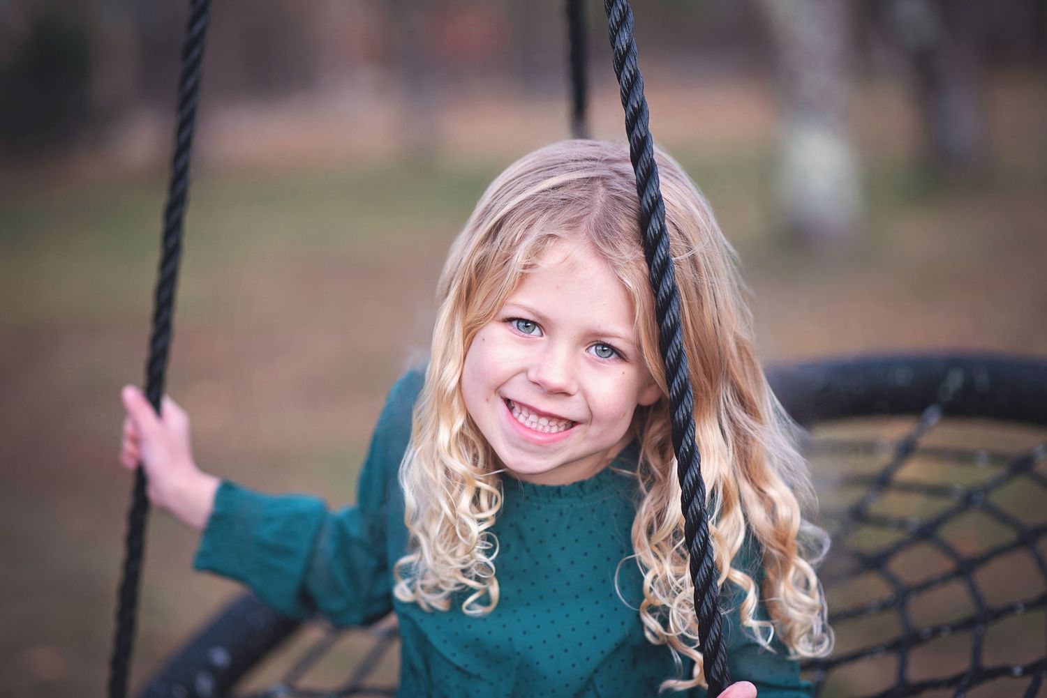 Child with curly blonde hair wearing a green sweater smiles while playing on a rope swing outdoors.