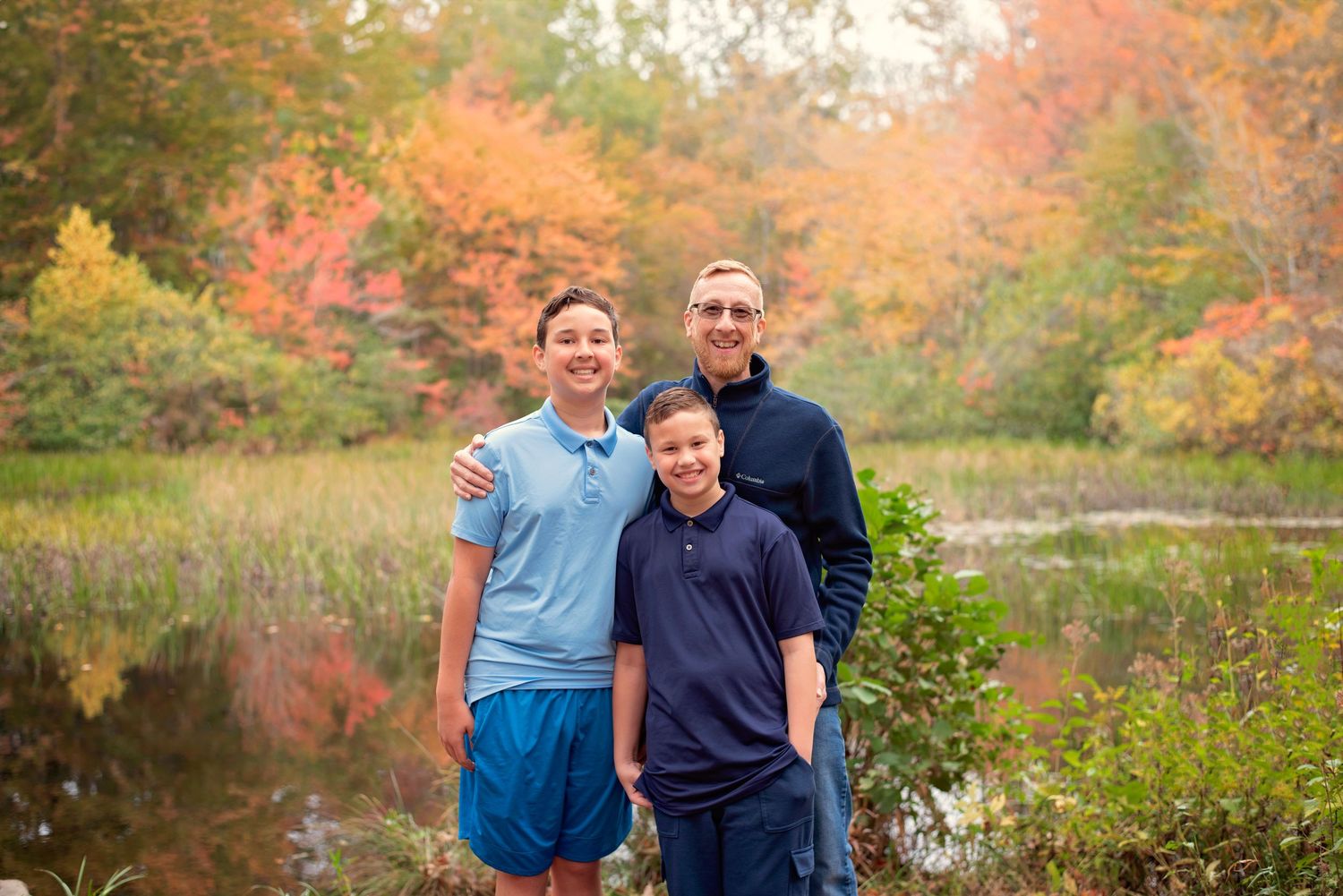 Three people stand together outdoors against a stunning backdrop of autumn foliage and a peaceful pond.