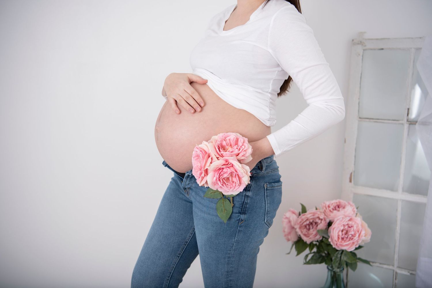 A pregnant woman in jeans and white shirt holds pink roses against her baby bump in a bright interior setting.
