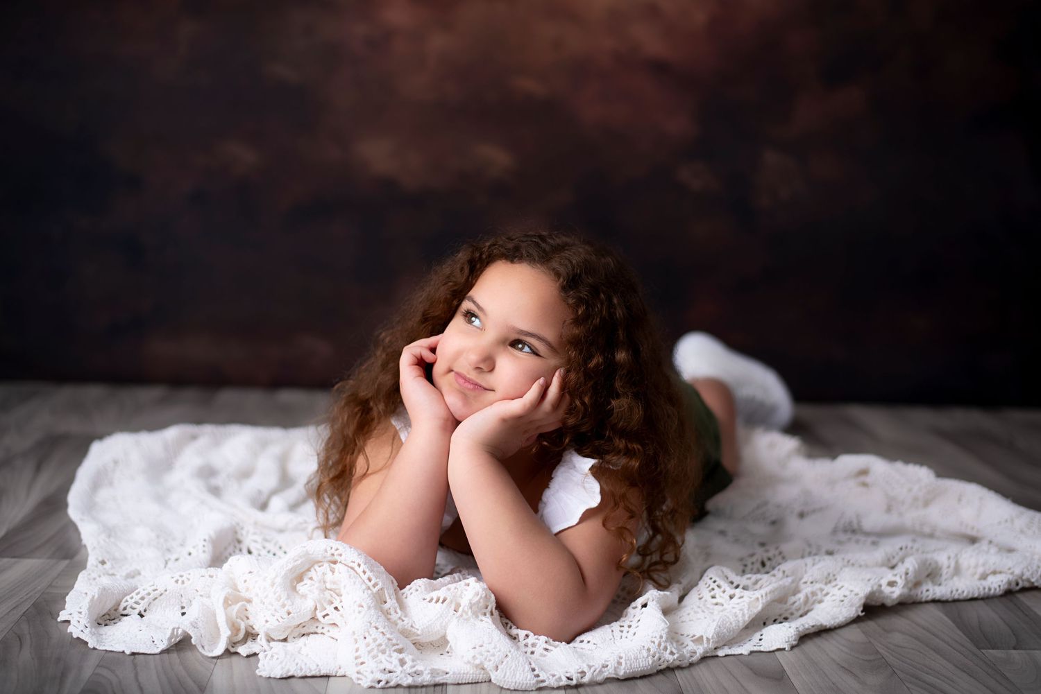 A young child with curly hair lies on white lace fabric while resting their chin on their hands against a dark backdrop.