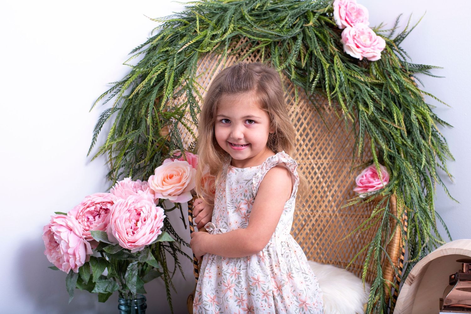 A small child stands in front of a floral greenery arch with pink peonies in a white lace dress.