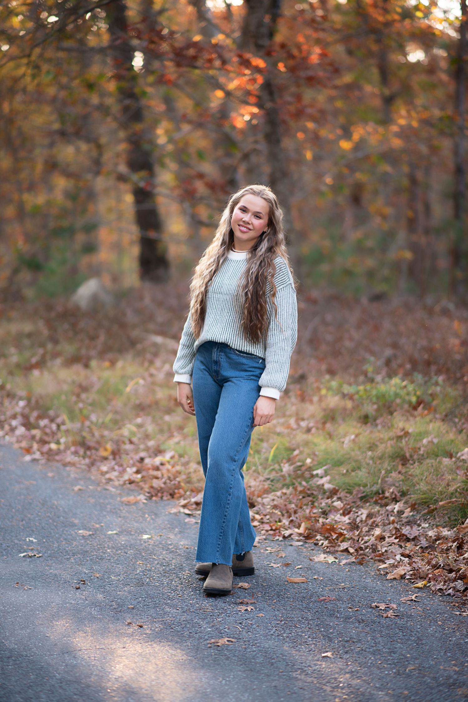 Autumn outdoor portrait showing someone in a cozy sweater and jeans standing on a path surrounded by fall foliage by RI Photographer.